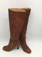 Pair of cognac leather knee high boots with side zippers and buckles, viewed standing upright from front and side angles. Visible leather texture and moderate wear marks.