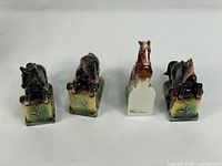 Four ceramic horse figurines arranged in a row, showing front view: two black horses on green-brown glazed bases, two brown horses, one with visible Lipper & Mann mark on base.