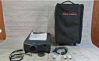 Mitsubishi LVP-X200A projector with power cord, cables, and user manual laid out on floor, with black carrying case behind
