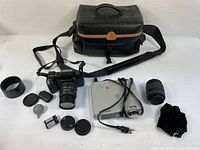 Full overhead view of Pentax SF10 camera body with Rokunar lens, additional Rokunar zoom lens, camera accessories including lens caps, lens hood, film canisters, blower, black camera bag with tan accent and strap, ViewSonic PJ250 projector with cables and remote on a white surface.