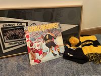 Photo showing Bruins scarf, knit hat with pompom, vinyl album, and framed Stanley Cup Champions banner.