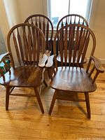 All four wooden dining chairs arranged two front and two behind near window light. Two captain chairs with armrests in front, two armless dining chairs behind. Medium brown finish, visible natural wood grain.
