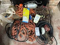 Photo showing assortment of heavy duty extension cords in orange, black, and brown, lamp kit parts in original packaging, electrical socket adapter, and various cables coiled together.