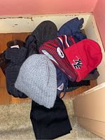 Pile of men's hats with black gloves on bottom. Hats include gray knit beanie, red hat with NE patch, and blue with red and white knit Patriots beanie.
