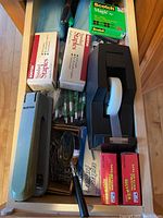 Photo of an open drawer containing various office supplies: boxes of staples, rolls of tape, a tape dispenser, and pens.
