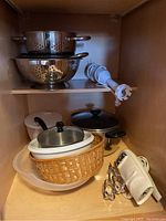 Full view of all kitchen items inside a wooden cabinet showing mixer, pots, colanders, bowls, beaters and chopper.