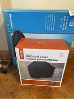 Blue boxed Brookstone massaging seat topper behind a boxed Classic Accessories BBQ grill cover for size 52 grills on carpet.