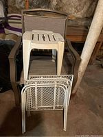 View showing two brown outdoor armchairs, one white plastic square side table placed on one chair, and three white metal mesh side tables (foldable and flat top) stacked and placed in front.