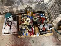 Wide view of assorted painting supplies including mineral spirits, paint cans, brushes, spray paint, tapes, and other items in boxes and on pallet on floor.
