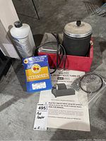 Photo showing the rotary rock tumbler, burnishing compound bottle, grit pack accessories, wire mesh, metal shot, and instruction manual on concrete floor.