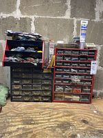 Two plastic storage organizer units on a wooden surface against a cinder block wall, with various fasteners visible in drawers and on top.