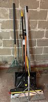 Full view of the lot showing a green metal leaf rake, black plastic snow shovel, roof rake with aluminum handle, and floor squeegee with yellow handle leaning against a brick wall.