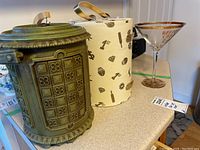Wide photo showing a green vintage plastic ice cube holder, a white bar supply container with black icons, and a gold-rimmed martini glass