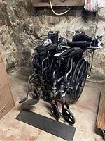 Two folded manual wheelchairs placed side by side against a stone wall, showing armrest controls and basket on one wheelchair.