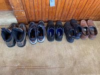 Wide side view of five pairs of men's shoes lined up against a wooden wall - includes brown/black Wolverine hiking boots, black/gray New Balance shoes, black and dark casual shoes, and brown slip-on Nunn Bush shoes