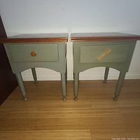 Front view showing two green painted solid wood night tables with wooden tops, each with one drawer, one drawer missing handle with tape on it.
