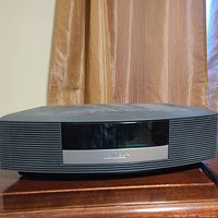Photo showing front and top of Bose Wave Radio in black, placed on wooden surface with a curtain background.