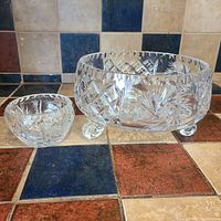 Two Bohemian crystal bowls placed side by side on a tiled surface with blue and beige square tiles. The larger bowl has three rounded feet and intricate cut crystal design featuring pinwheels and crosshatch pattern.