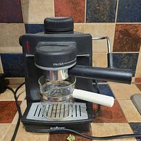Black Salton espresso machine on tiled kitchen counter with glass pot, dial marked 'UNLOCK', and steam wand.