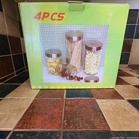 Photo showing box packaging of 4 clear glass storage canisters with metal lids, featuring images of various sized jars with metal lids on the front.