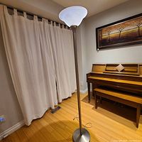 Tall floor lamp with frosted white glass upward bowl-shaped shade, metallic stand and base, set in a room with hardwood floor and piano.