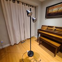 Full view of the black floor lamp with two metal cone-shaped adjustable shades on single pole, placed on a hardwood floor in a room with a piano and a painting on the wall.