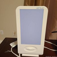 Verilux light therapy lamp with white frame and large rectangular light panel, placed on a dark table with power cord and a control dial on front