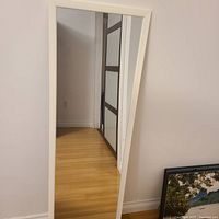 Full view of the Ikea standing mirror with white frame leaning against the wall on wood floor.
