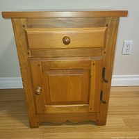 Front view of solid oak night table showing drawer, cabinet door with wooden knobs and black metal hinges.