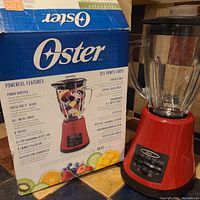 Red Oster blender with glass jar placed on tiled kitchen counter next to its box showing features and specs