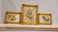 Photo showing set of 3 watercolor floral paintings framed in gold ornate frames arranged on a shelf.