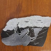 Stone carving showing two Inuit hunters with harpoons dressed in parkas, hunting seals by rocky shore with mountains and a cloudy sky in the background. Signed 'Iguk'.