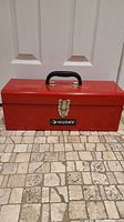 Photo of red Husky toolbox placed on tiled floor in front of door, showing front and top handles and latch.