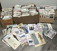 Wide view of multiple boxes and stacks containing numerous vintage sewing pattern envelopes from various well-known brands and designers.