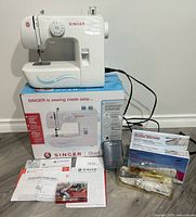 Singer Start 1304 white automatic sewing machine placed on its original box. Small sewing accessories including a foot pedal, manuals, and a case of bobbins and threads arranged in front. Battery operated Narada handheld sewing machine in box.