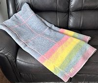Folded wool blanket laid on a brown leather chair showing light blue side with pink grid lines and rainbow stripe edge