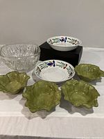 Full lot of seven bowls on white surface against gray wall: one large clear glass, two floral ceramic, four green ceramic bowls.