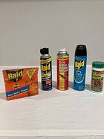 Photo showing five different bug repellent items arranged side by side on a white surface - two Raid spray cans, one Do It Best spray can, Raid Deep Reach Fogger box, and Shake Away powder container.