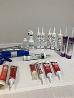 Overall items on white surface showing various caulking and sealant tubes with spray bottles and 3 caulk guns included.