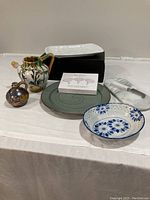 Overview of lot showing all dishes and items including asparagus plate, green plate, blue floral dish, cheese plate with knife, yellow and green pitcher, and boxed porcelain cheese marker set