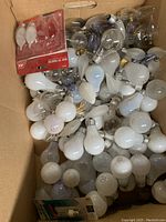 Multiple incandescent and other light bulbs of different sizes and shapes piled in a box, including some in original packaging.