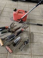 Garden hand tools including looper shears, bulb planters with wooden handles, hand clippers, and a red gas can on a concrete surface.