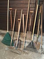 Photo of 11 assorted garden tools including 3 shovels, 3 lawn rakes, 2 metal garden rakes, cultivator, hoe, pitchfork and sledgehammer leaning against a brown wall.