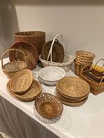 Collection of various woven baskets of different sizes and colors arranged on floor against wall, showing diversity in handle types and basket shapes.