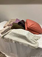 Assorted blankets and pillows displayed on a white surface with different textures and colors including purple/beige wool, military green, and plaid patterns along with red pillows.
