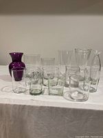 Photo showing 12 vases in various shapes and sizes, including one purple opaque vase and several clear glass vases in cylindrical, square, and rounded shapes.