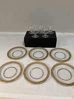 Six gold-rimmed glass plates arranged on a white surface with five crystal sherbet dishes elevated on a black box behind them.