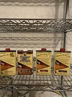 Four 16 oz containers of Howard Restore-A-Finish wood refinishing liquids in Maple Pine, Dark Oak, Walnut, and Cherry on a metal shelf.