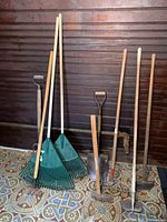 Full view of 9 assorted lawn and garden tools against dark wooden wall showing rakes, shovel, hoe, cultivator, pitchfork, sledgehammer, and edger.