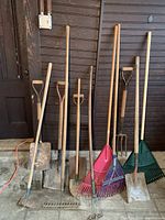 Photo showing eleven assorted lawn and garden tools including shovels, plastic and metal rakes, hoe, cultivator, pitchfork, and small spade propped against a dark wood wall.